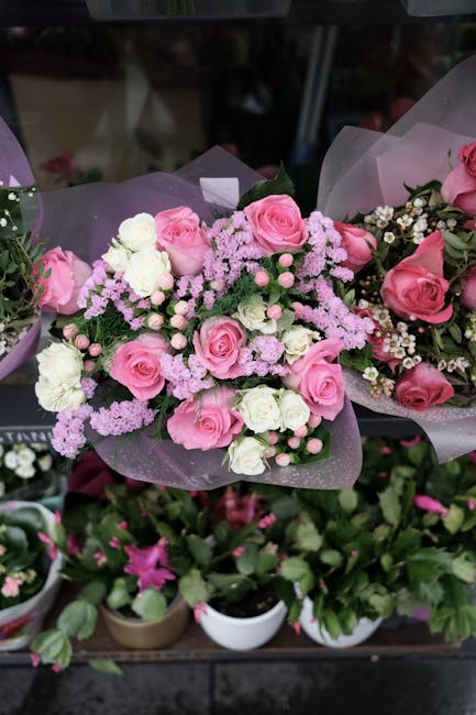 A floral arrangement featuring a bouquet of fresh pink roses, white carnations, and small pink and white filler flowers, all delicately wrapped in purple tissue paper. The bouquet is arranged with gre