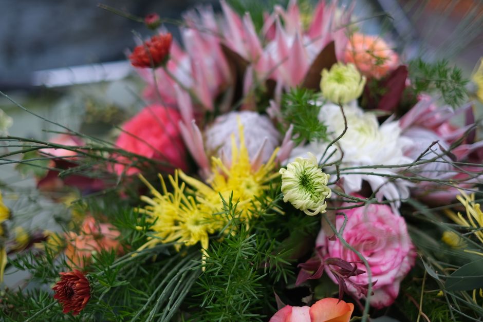 A vibrant floral arrangement featuring a variety of fresh flowers including soft pink roses, greenish-white ranunculus, and yellow spider chrysanthemums, accented with dark red blooms and wispy greene