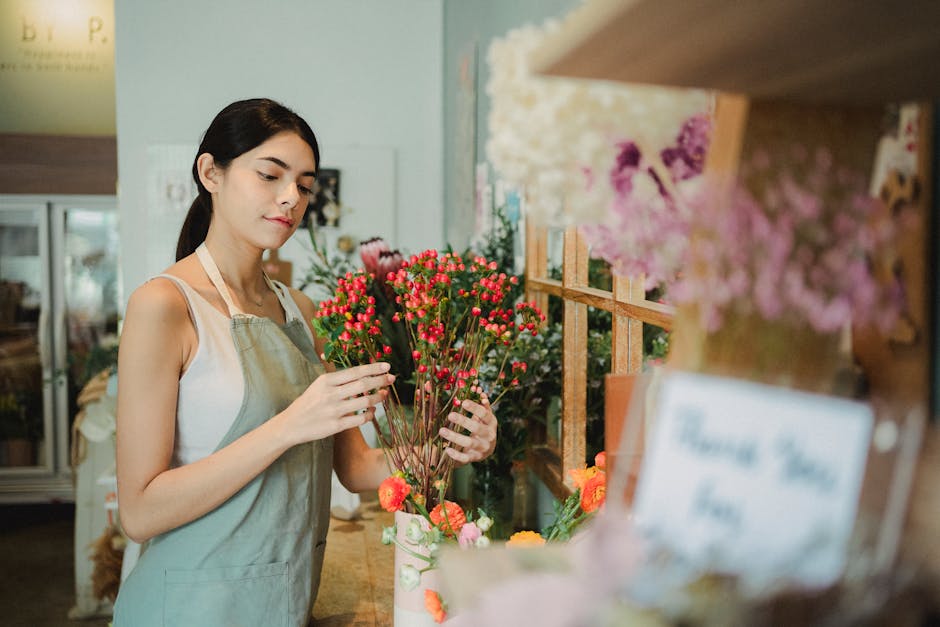 A young female florist with dark hair tied back, wearing a light-colored apron over a sleeveless top, arranging a bouquet of small, round, vivid pink flowers with green foliage. She stands behind a wo