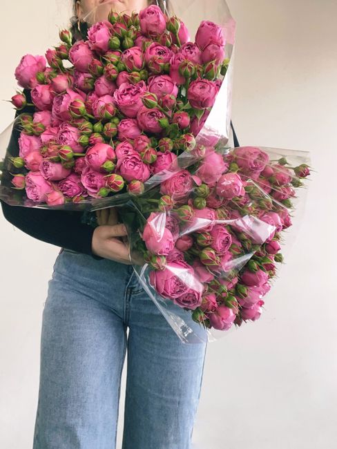A person holding a large bouquet of fresh pink roses with numerous tightly closed buds and green stems, wrapped in clear plastic with a pink ribbon. The bouquet is accented with dark green foliage, an