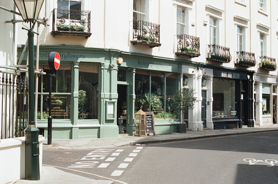 A quaint florist storefront on a quiet street in Parsons Green, London, painted in a sage green color with large glass windows displaying fresh flowers inside. The shop features black wrought iron bal